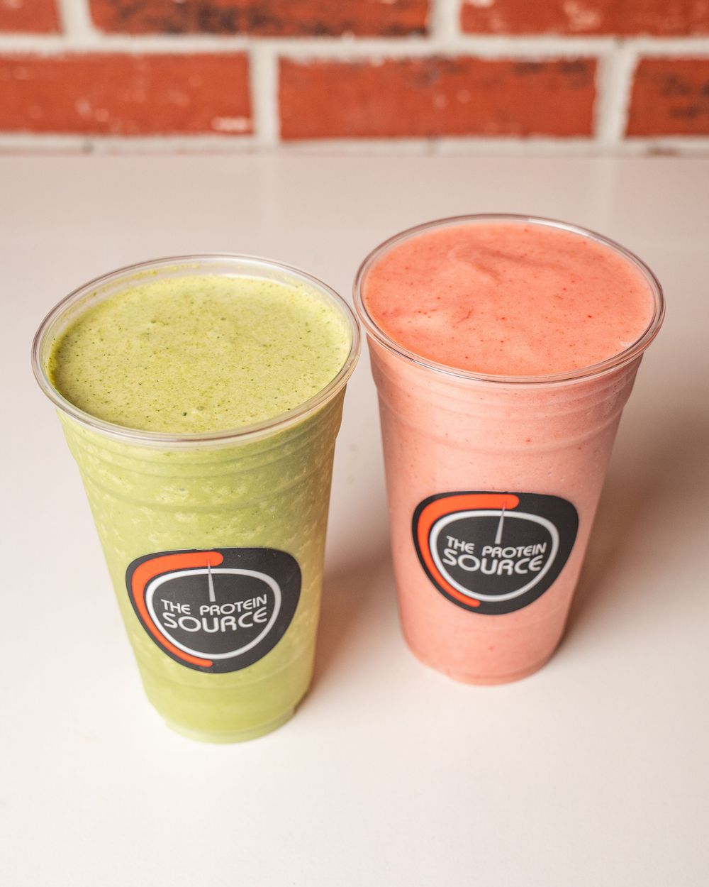 Two colorful smoothies from The Protein Source, one green and one pink, served in clear cups on a white surface.