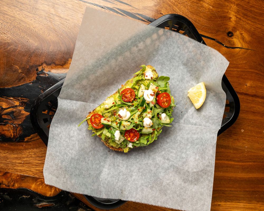 Avocado toast topped with cherry tomatoes, greens, and feta on parchment paper with a lemon wedge.
