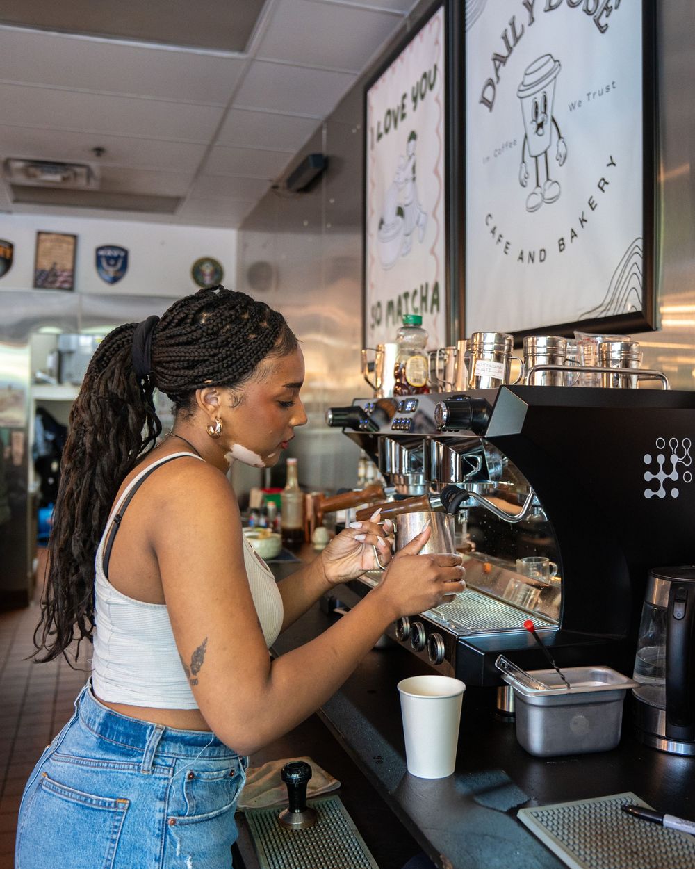 A barista in a white crop top and jeans operates an espresso machine at Daily Dose Cafe and Bakery.