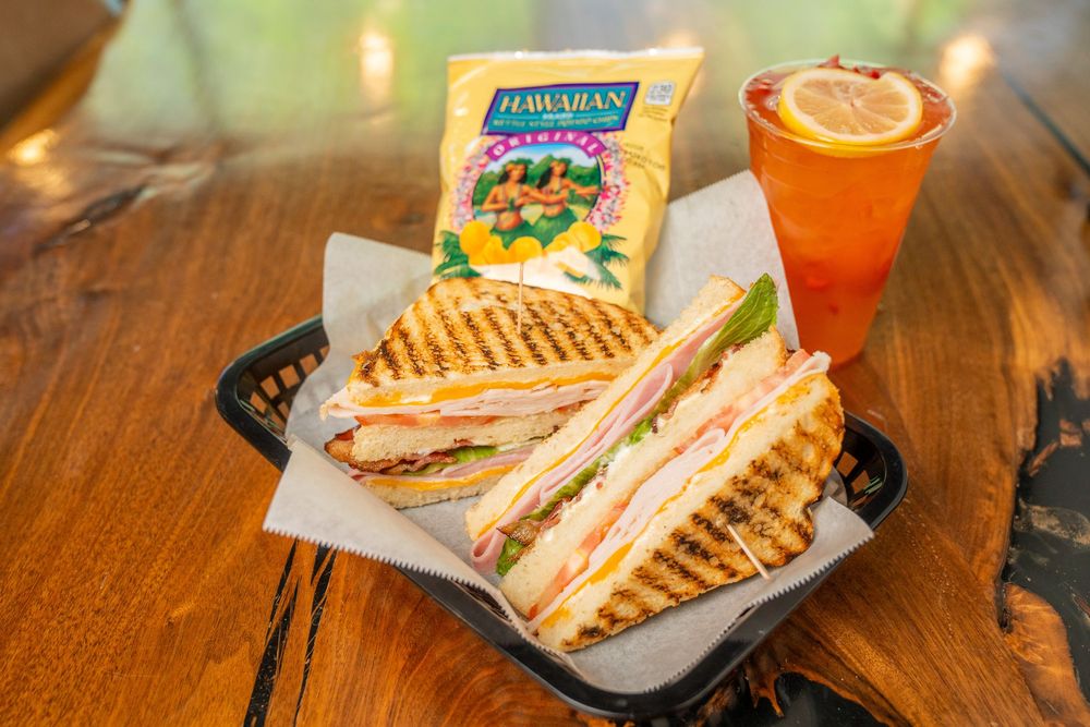 Grilled panini sandwich with ham and cheese, Hawaiian chips, and iced tea served in a black basket on a wooden table.