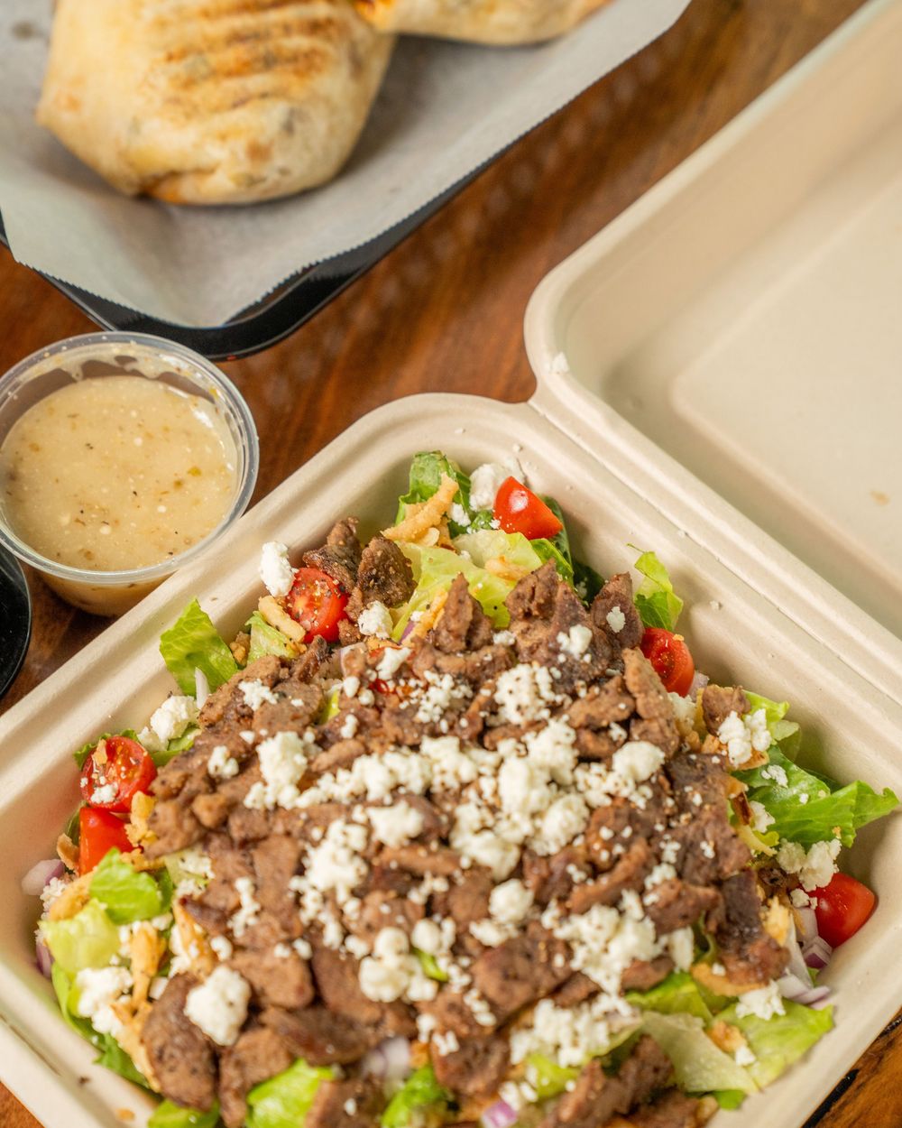 Fresh salad with seasoned meat, crumbled feta, tomatoes, and lettuce in a takeout container with dressing on the side.