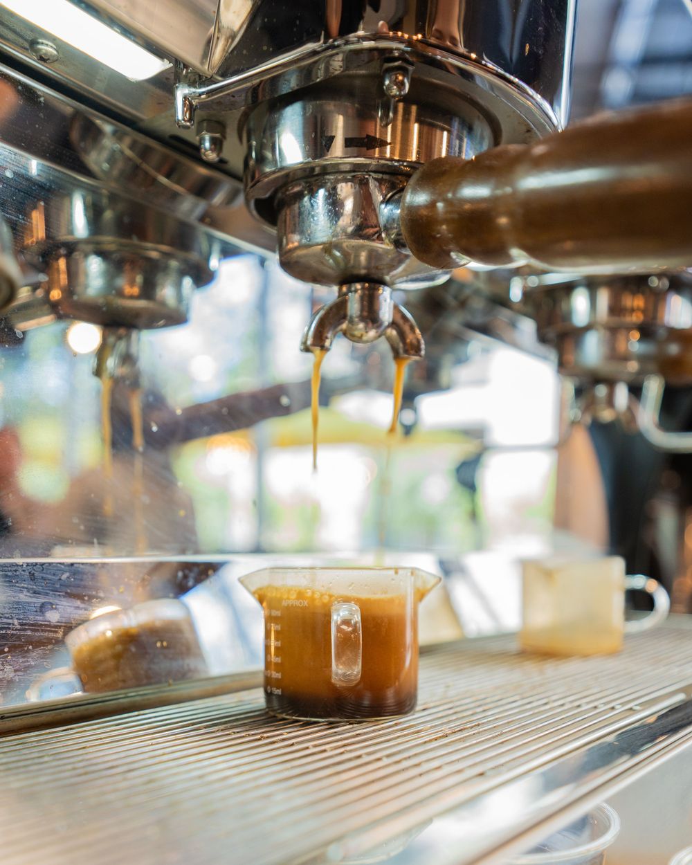 Espresso shot pouring from a professional chrome coffee machine into a small glass measuring cup on a metal drip tray.