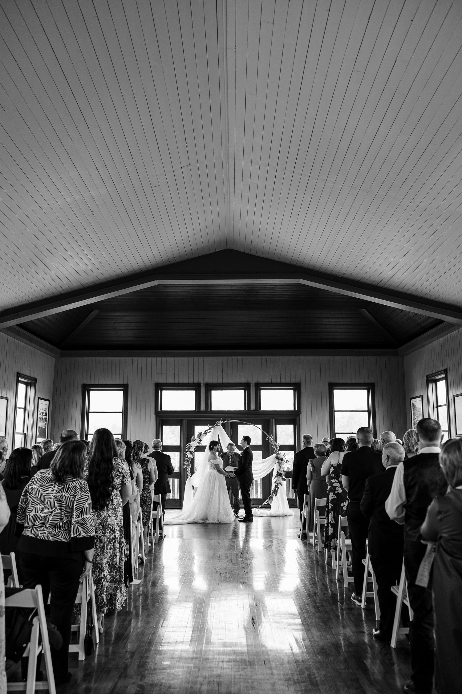 Black and white photo of a wedding ceremony in a bright barn venue with guests seated in white chairs along the aisle.