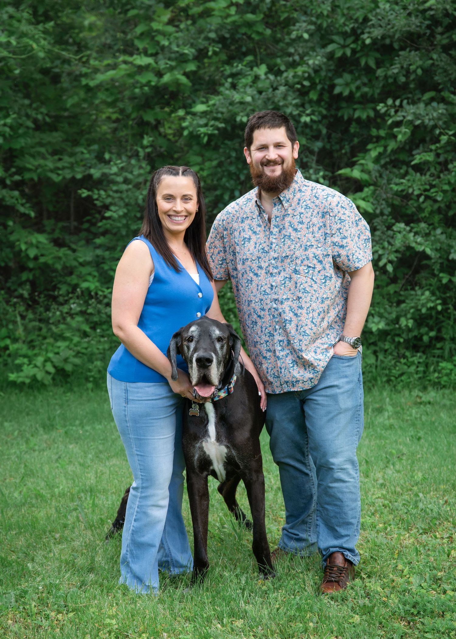A couple smiles outdoors with a large black Great Dane dog on a green lawn surrounded by trees.