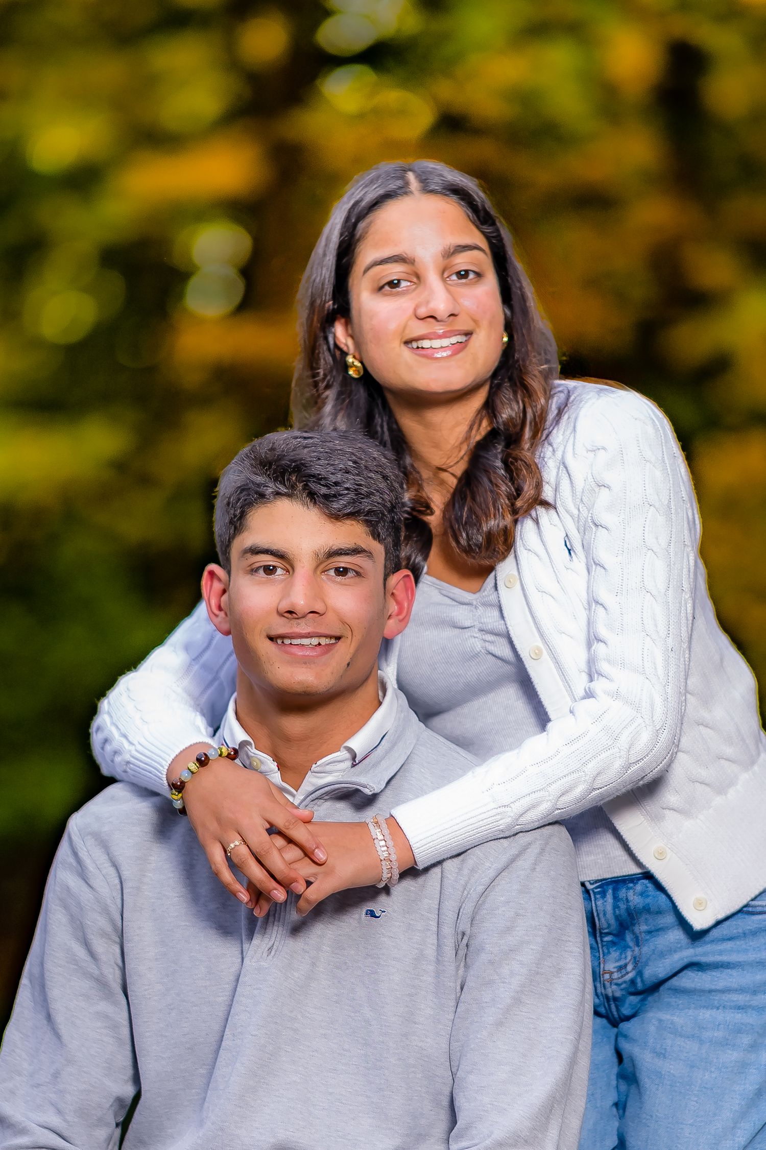 Two smiling young people pose together outdoors with colorful autumn foliage in the background.
