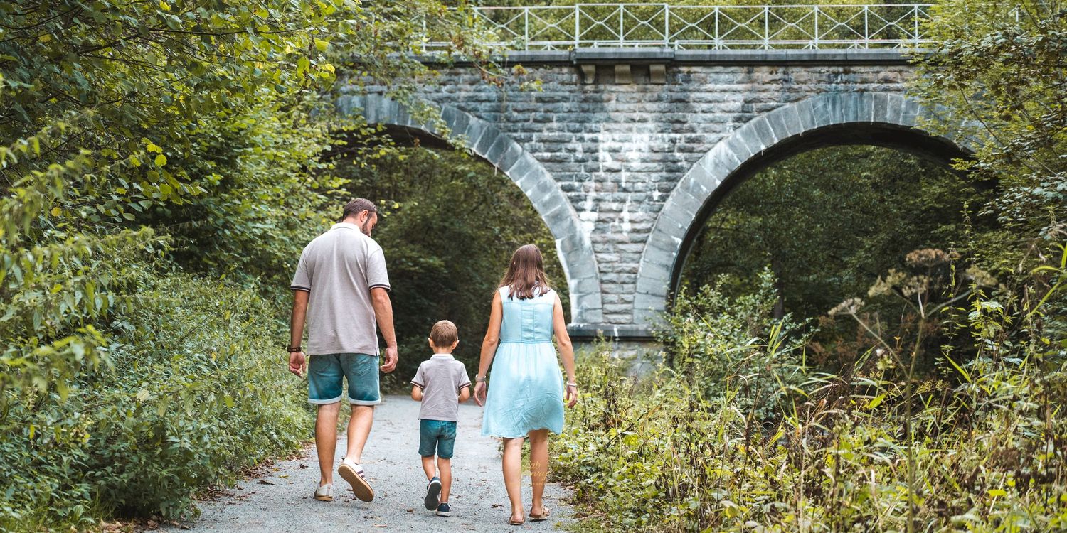 Famille sous une arche en pierre, chemin boisé (père, mère en robe bleue, enfant)