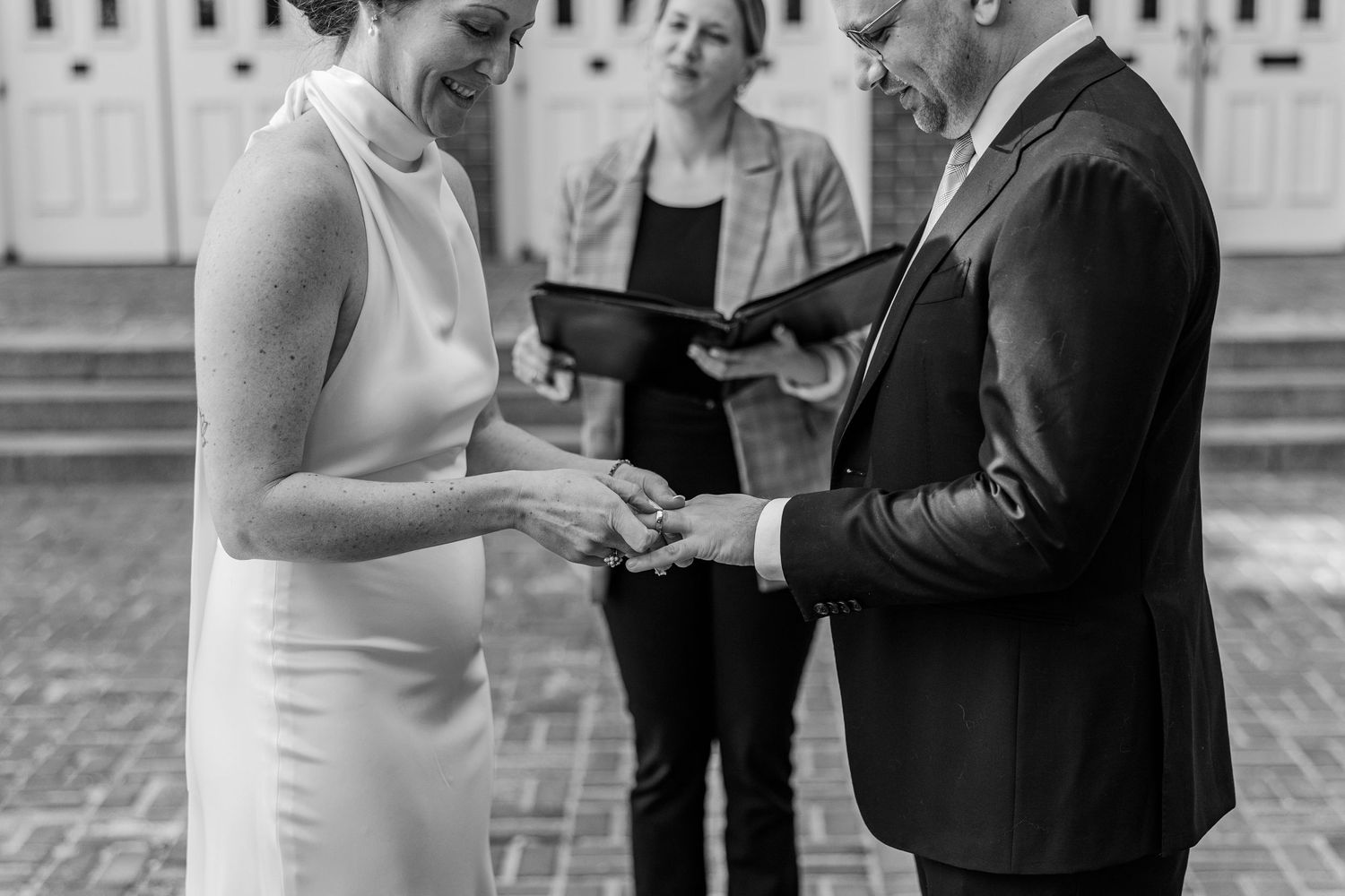 Black and white wedding ceremony photo of couple exchanging rings outdoors with officiant in background.