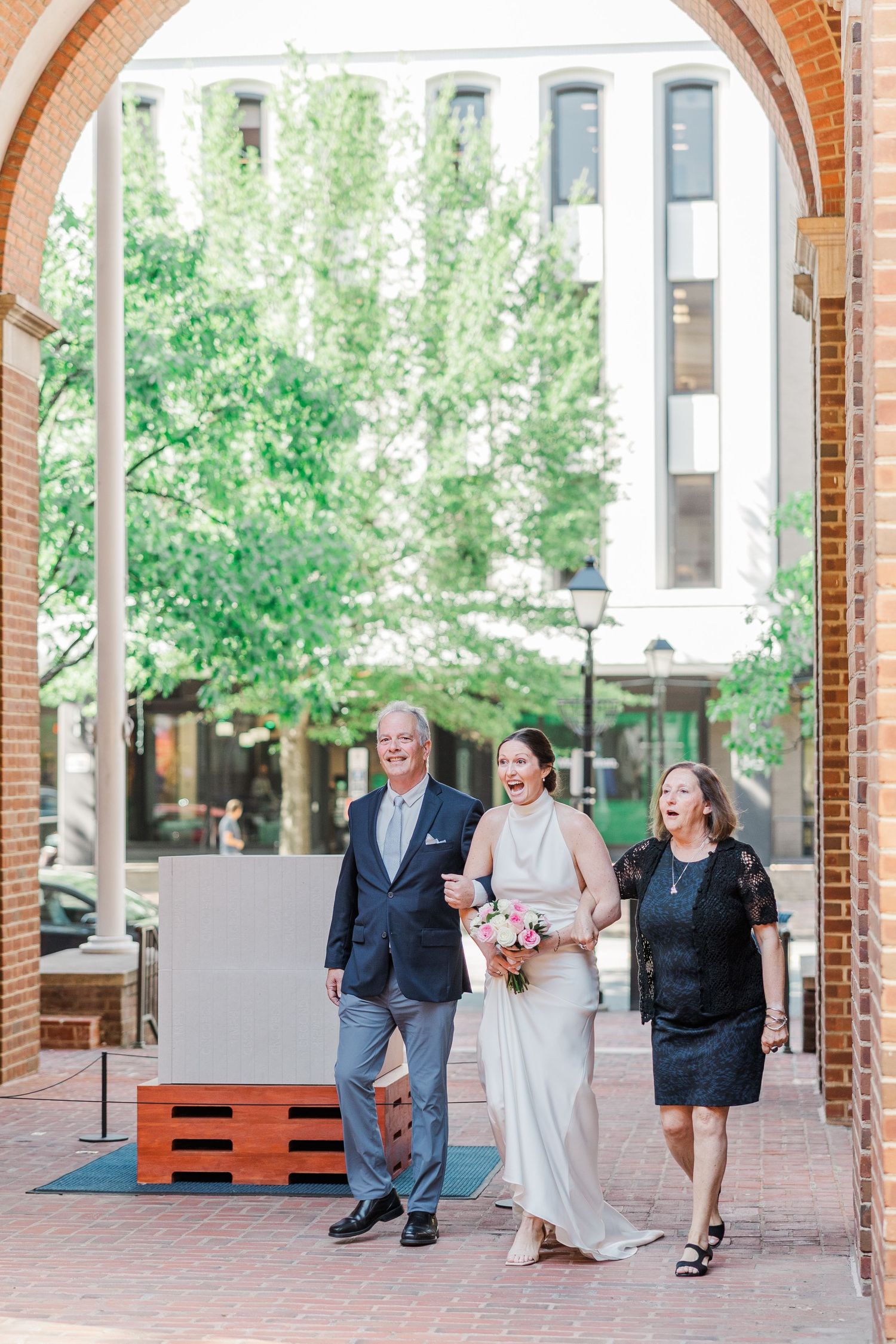 Bride walks with parents through brick archway in urban setting, all three laughing joyfully on wedding day.