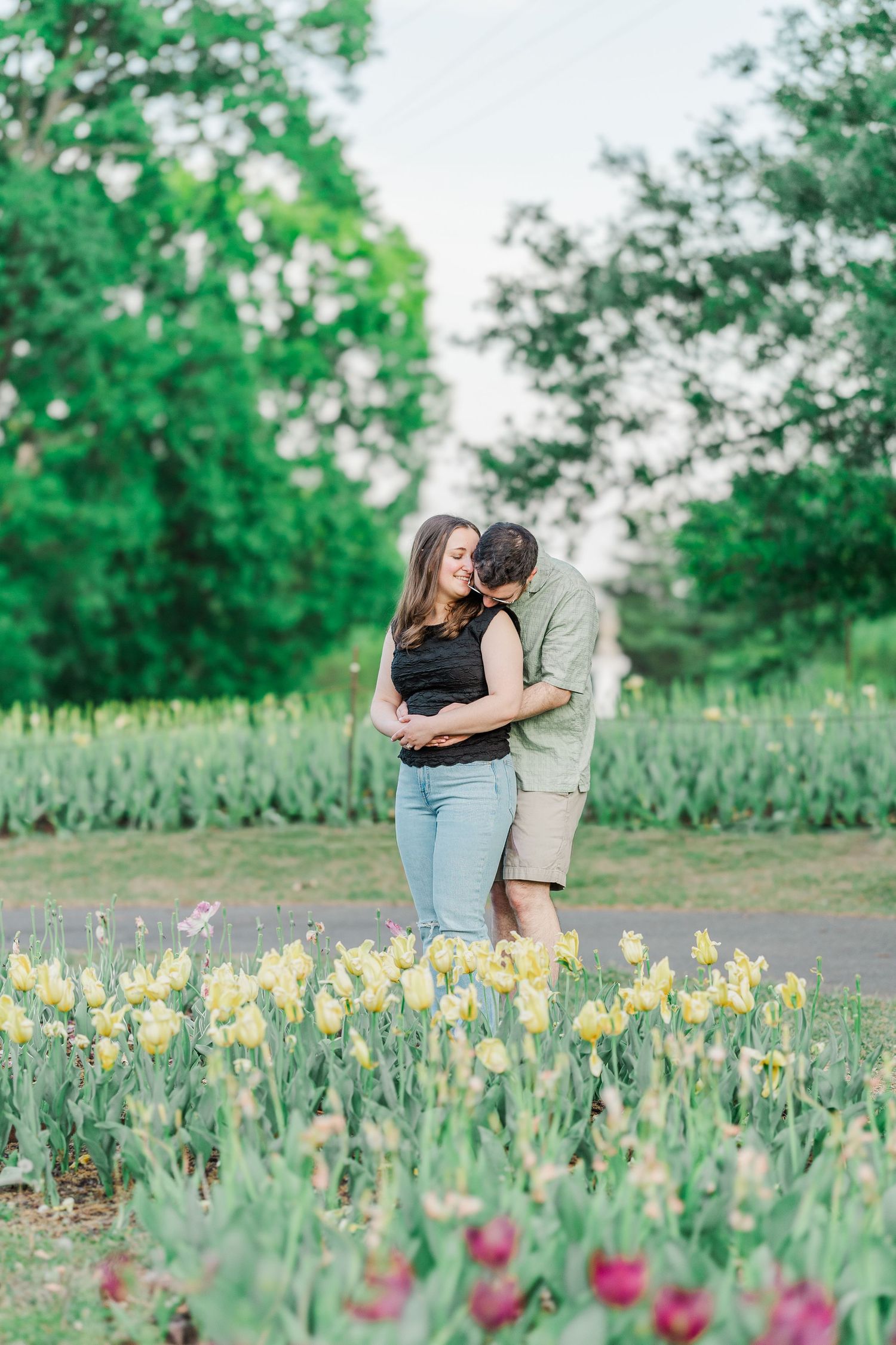 Couple embracing in a garden surrounded by blooming tulips and lush green trees during a romantic outdoor photoshoot.