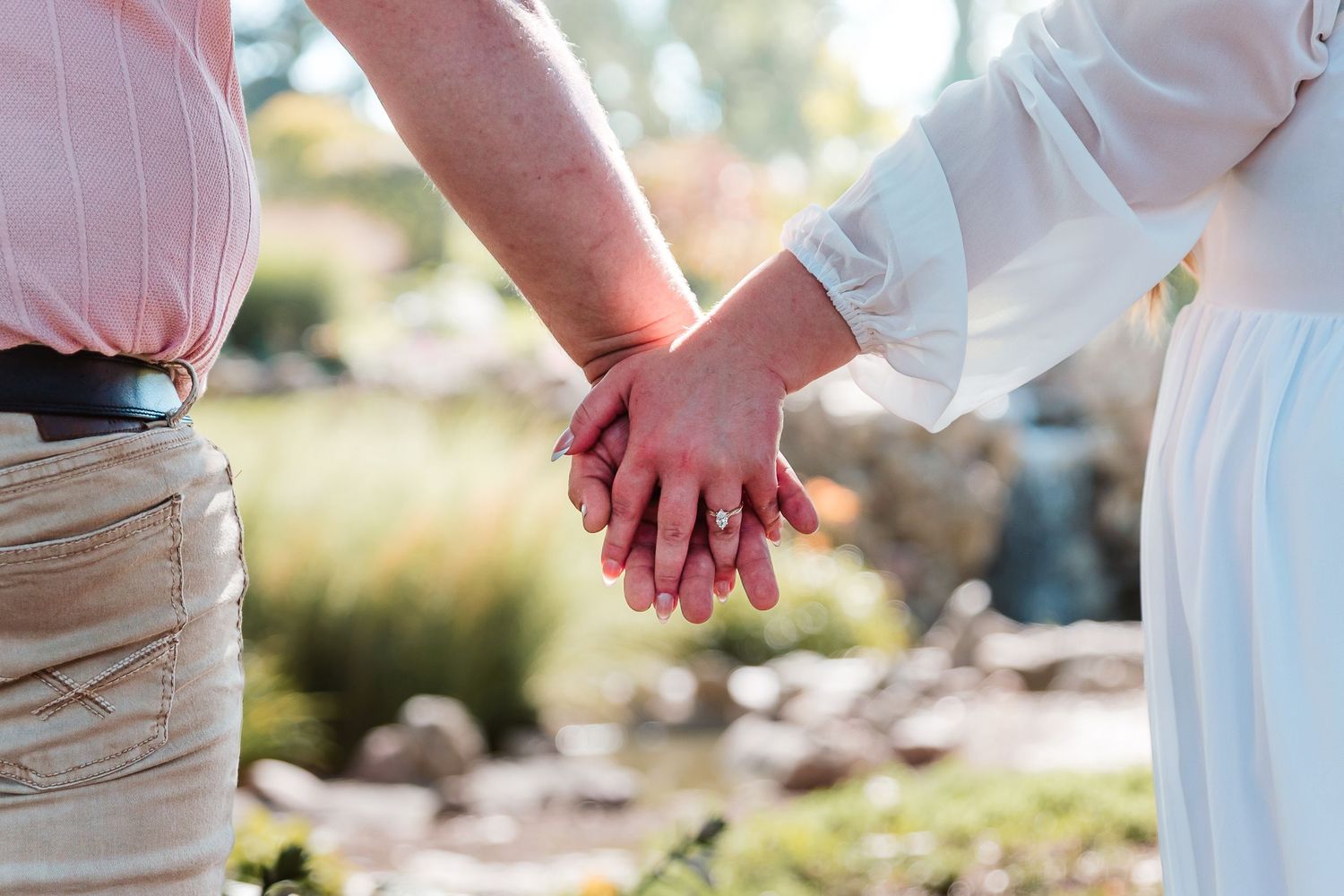 Couple holding hands outdoors, woman wearing engagement ring, romantic garden setting with soft bokeh background.