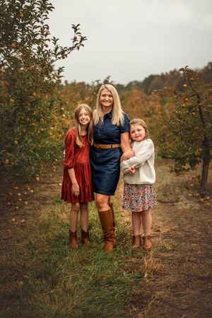 A woman and two girls pose in an apple orchard in fall, wearing dresses and boots.