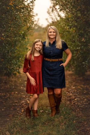 A smiling mother and daughter pose together in a tree-lined orchard, wearing coordinating fall dresses and boots.