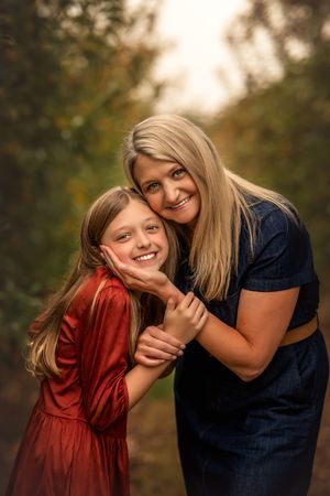 A blonde woman in navy dress hugs smiling girl in red velvet dress outdoors in autumn setting.