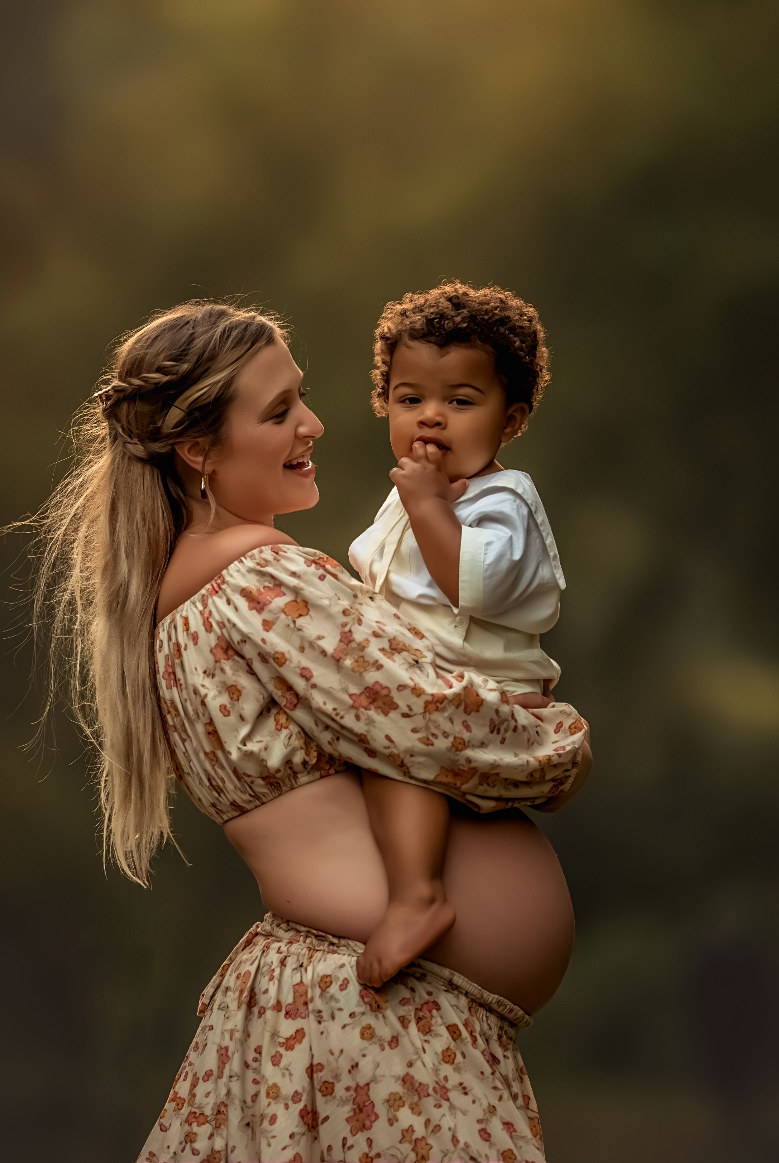 Pregnant woman in floral dress holding a toddler boy outdoors in warm golden light during a maternity photo session.