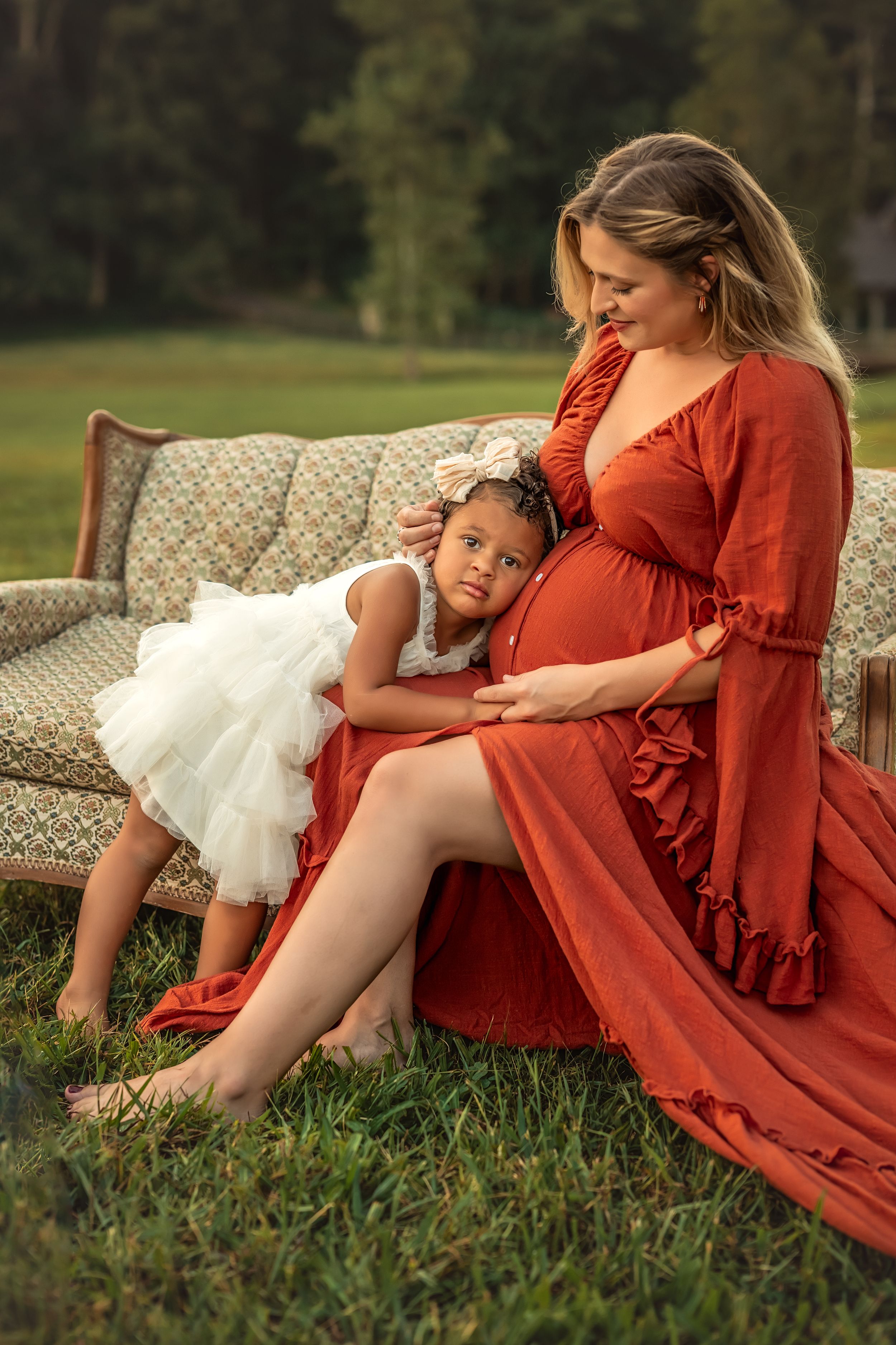 Pregnant woman in red dress sitting outdoors with young girl in white dress resting on her baby bump.