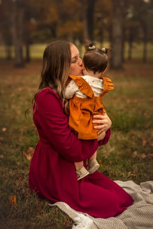 Mother in red dress kisses baby in orange outfit while sitting on blanket in autumn park.