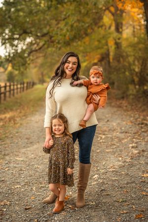 A mother in a cream sweater holds her baby girl while her toddler daughter stands beside her on a fall nature path.