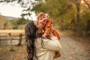 A dark-haired mother lovingly lifts her baby in an autumn outdoor setting with fall foliage.