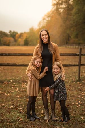 A mother smiles with her two young daughters hugging her in an autumn outdoor setting with fall foliage.