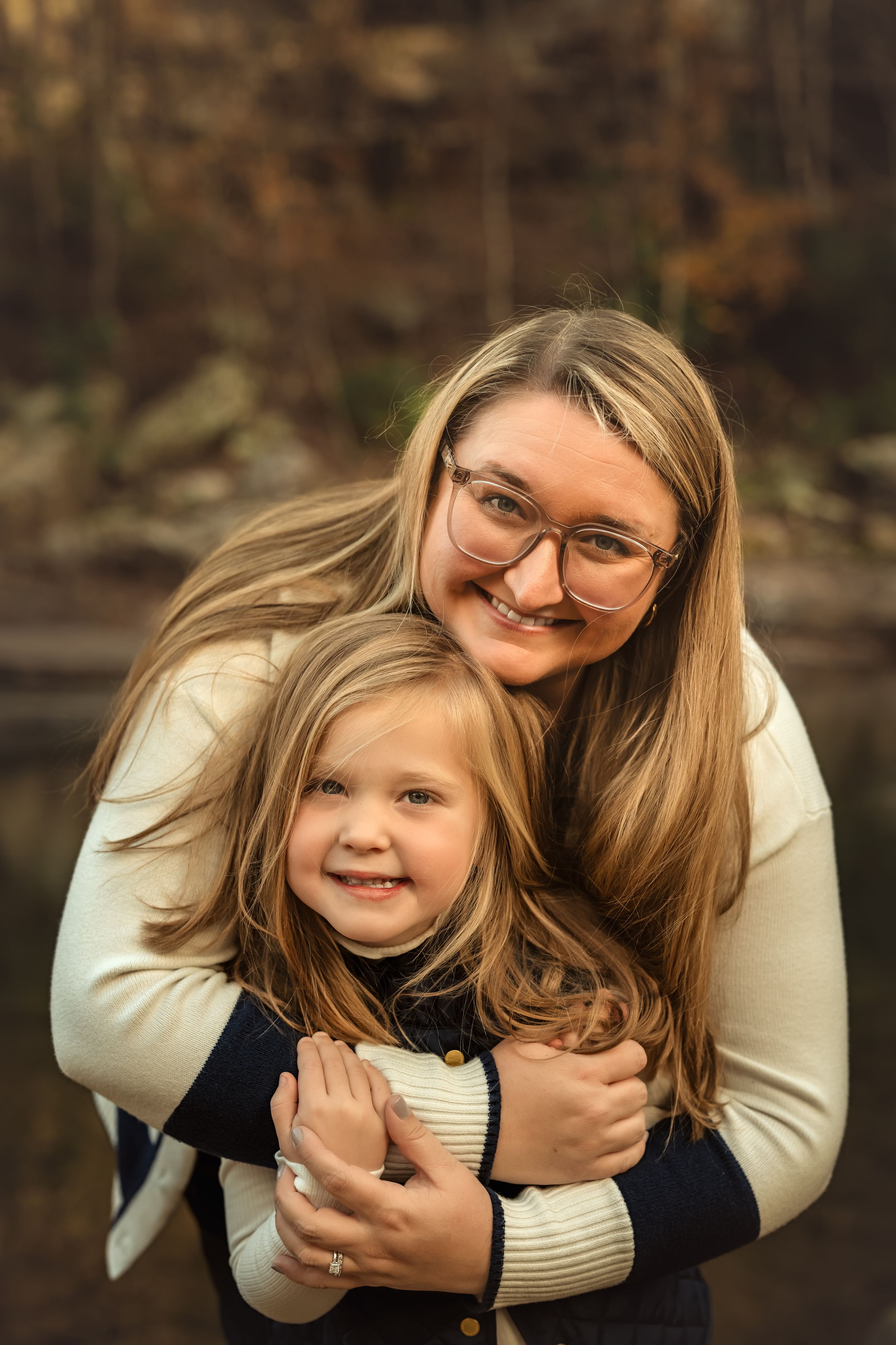A smiling woman with glasses hugs a young blonde girl outdoors in a warm autumn setting.