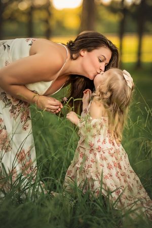 A mother kisses her young daughter in a sunlit green field, both wearing matching floral dresses.