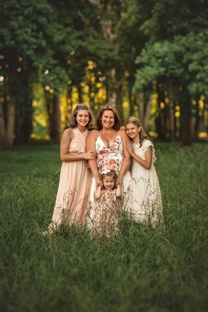 A mother and three daughters pose together in a lush green field wearing floral and pastel dresses.