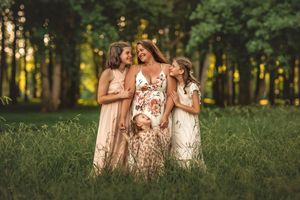 Pregnant woman in floral dress surrounded by three daughters in a sunlit green meadow with trees.