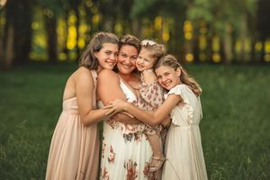 A mother and three daughters embrace outdoors in a sunlit park, wearing elegant dresses for a family portrait.