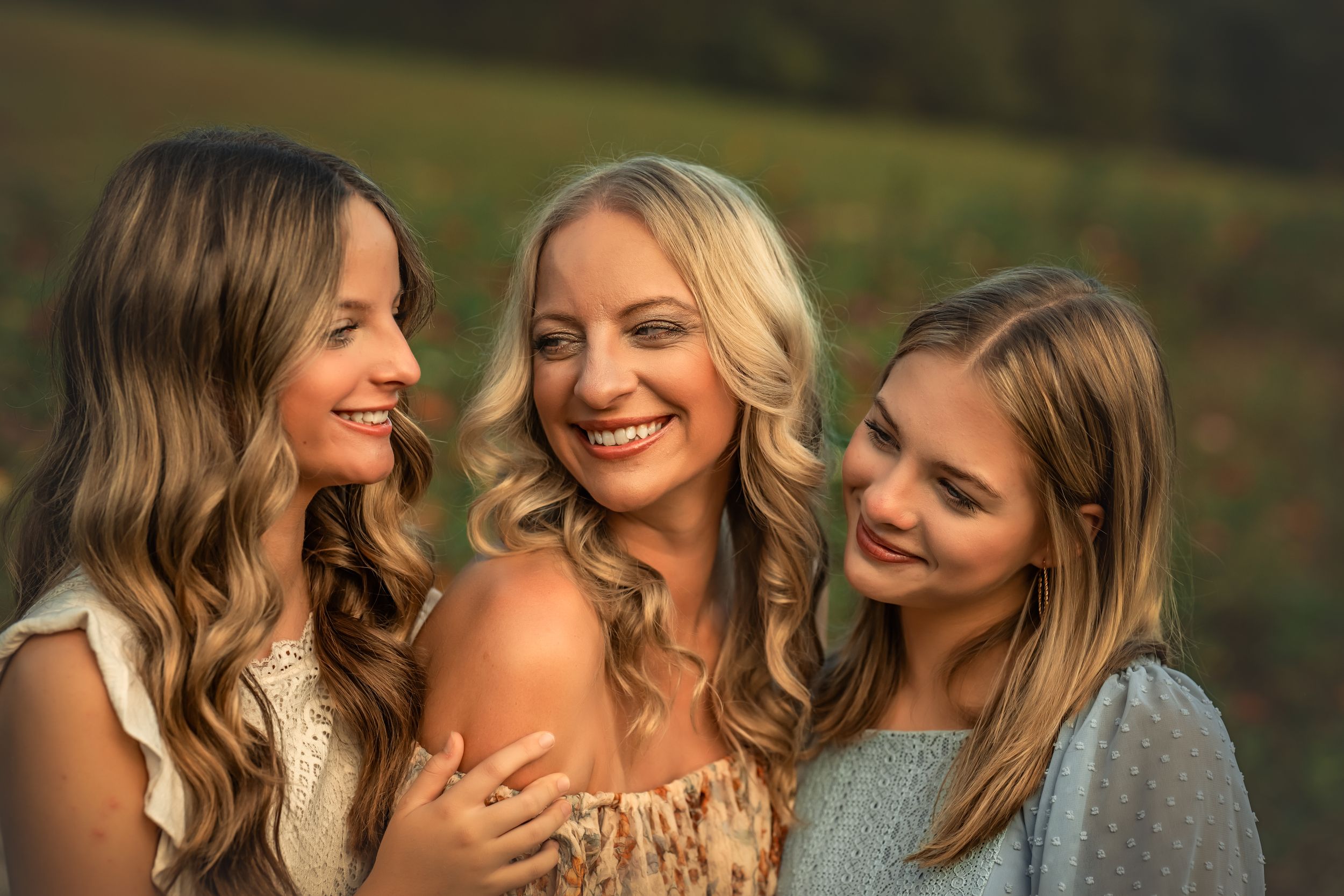 Three smiling young women with long hair pose together outdoors in warm golden sunlight in a green field.
