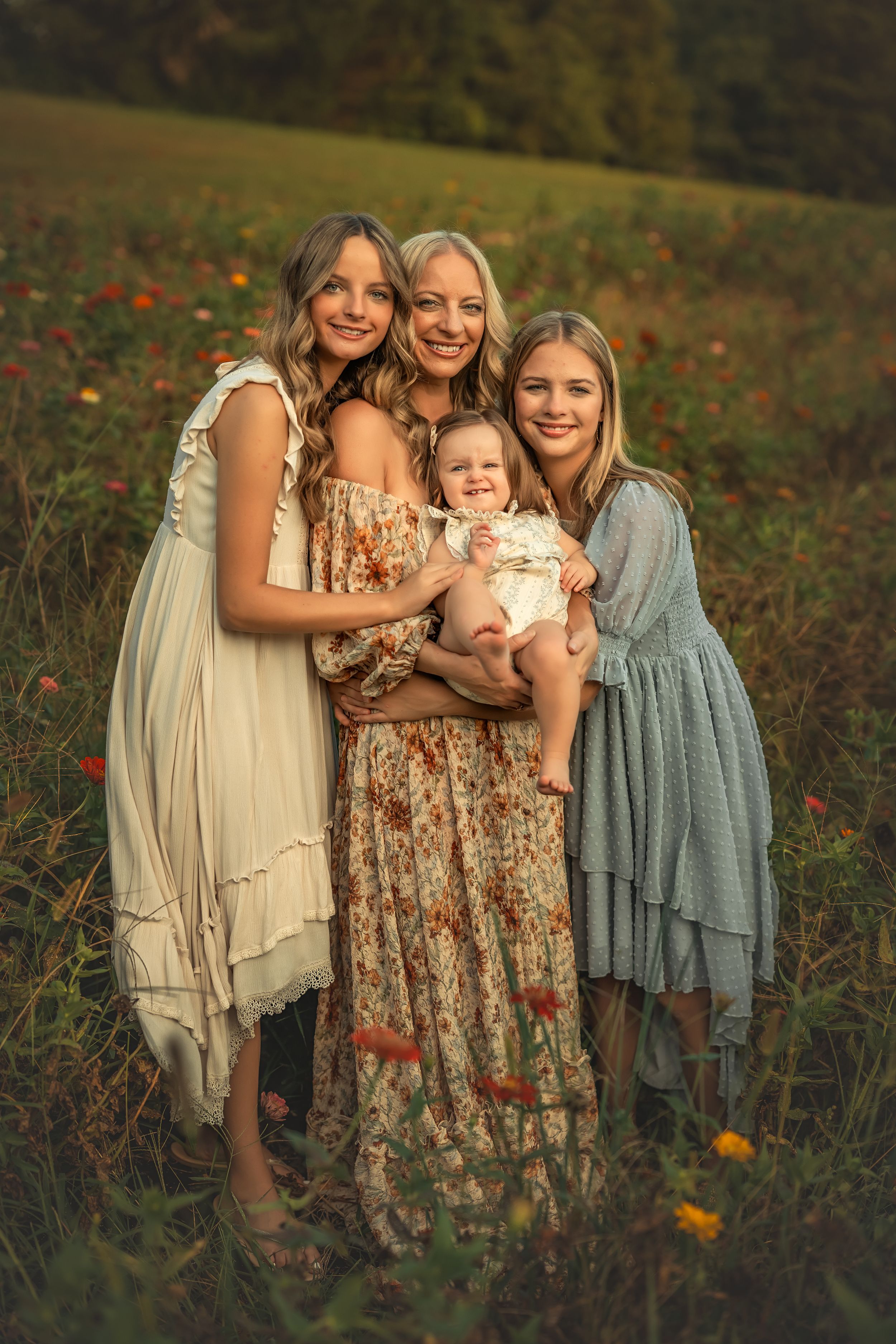Four generations of women pose in a sunlit flower field, wearing flowy dresses, with the eldest holding a baby.