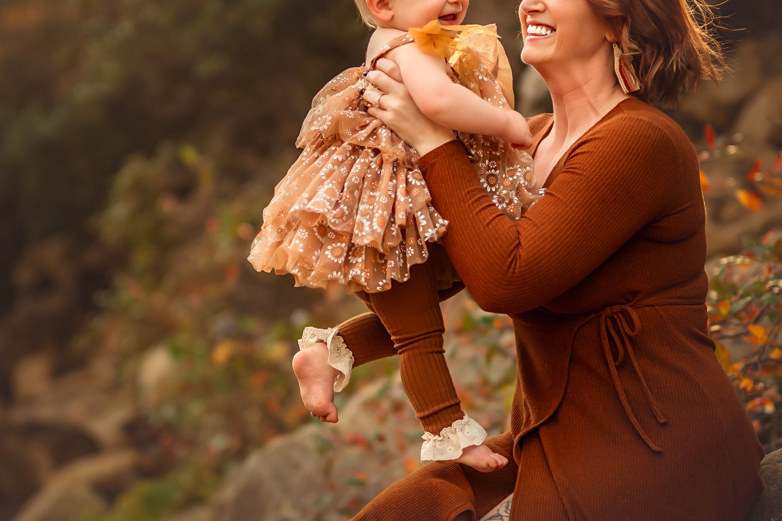 A joyful mother in a rust dress lifts her laughing baby girl in a floral outfit outdoors in autumn.