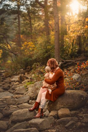 A mother hugs her young child on rocky terrain surrounded by autumn foliage in golden sunset light.
