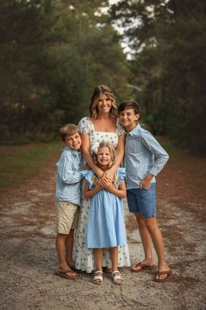 A smiling woman in a floral dress poses with three children on a tree-lined path during a family portrait session.
