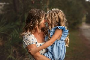 A mother holds her young daughter close, their foreheads touching tenderly outdoors in soft golden light.