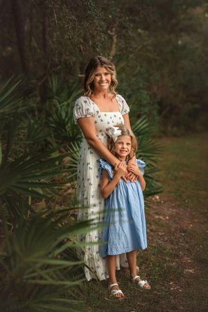 A smiling mother in a floral dress embraces her young daughter in a blue dress outdoors among lush green palms.