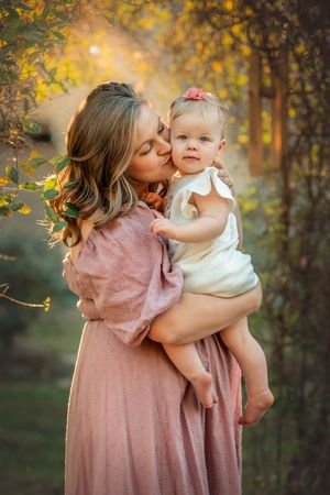 A mother in a pink dress kisses her baby girl in a white outfit outdoors in warm golden light.