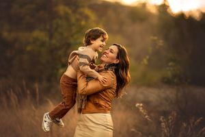 A mother joyfully lifts her young son in a sunlit autumn field, both laughing and sharing a tender moment.
