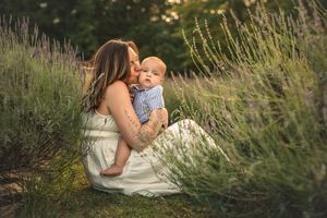 Mother kissing baby in lavender field at golden hour, wearing white dress outdoors.