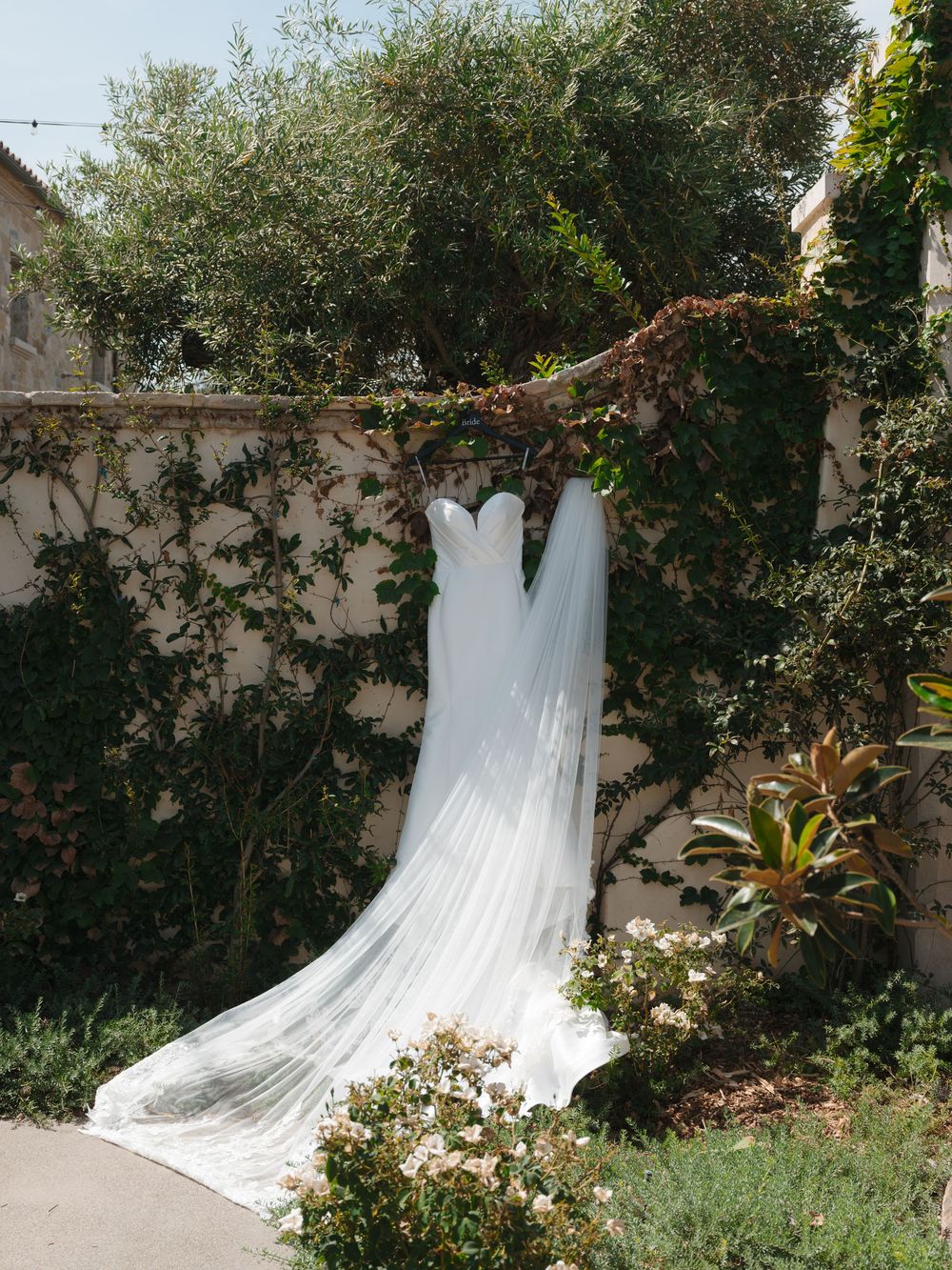 Elegant white wedding dress with long veil hanging from a tree branch against a garden wall.