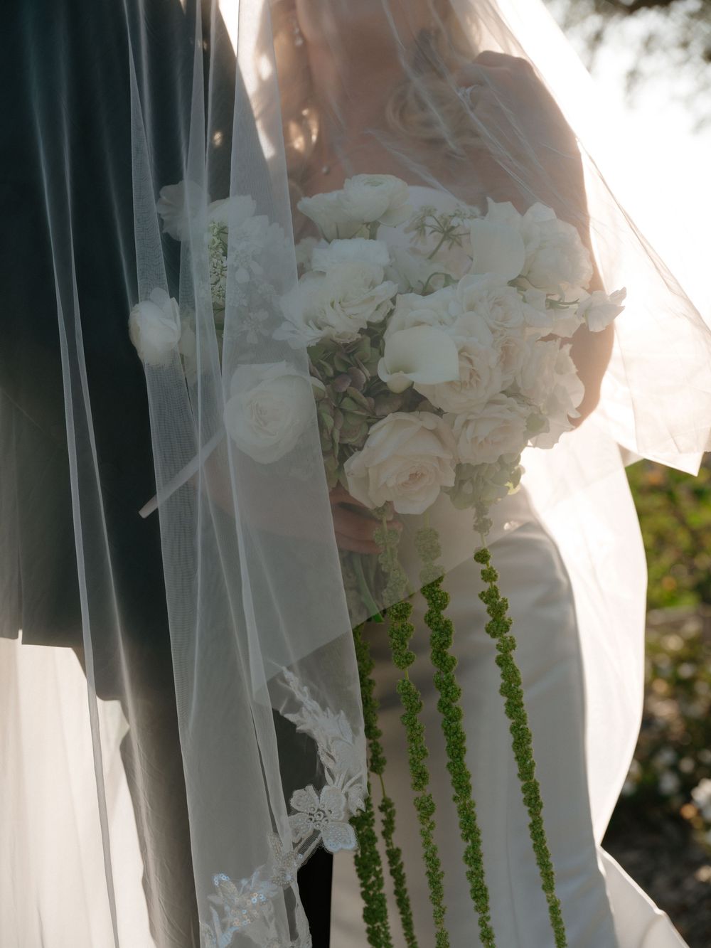 Bride holding white rose cascading bouquet with green accents, wearing lace-trimmed veil at outdoor wedding ceremony.
