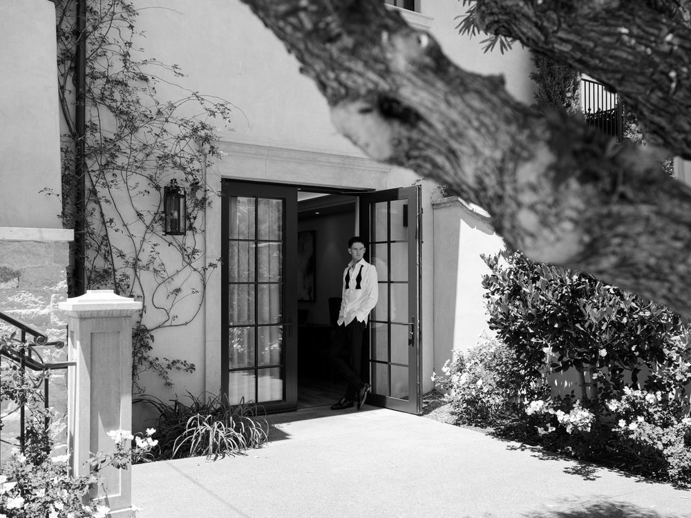 Black and white photo of a person standing in open French doors of an elegant building surrounded by lush greenery.