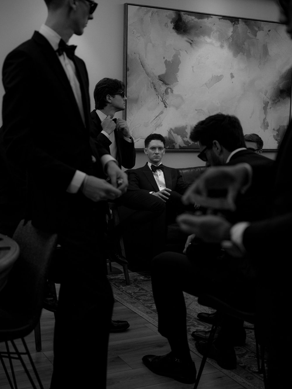 Black and white photo of men in tuxedos gathered in an elegant room with abstract art, exuding formal sophistication.