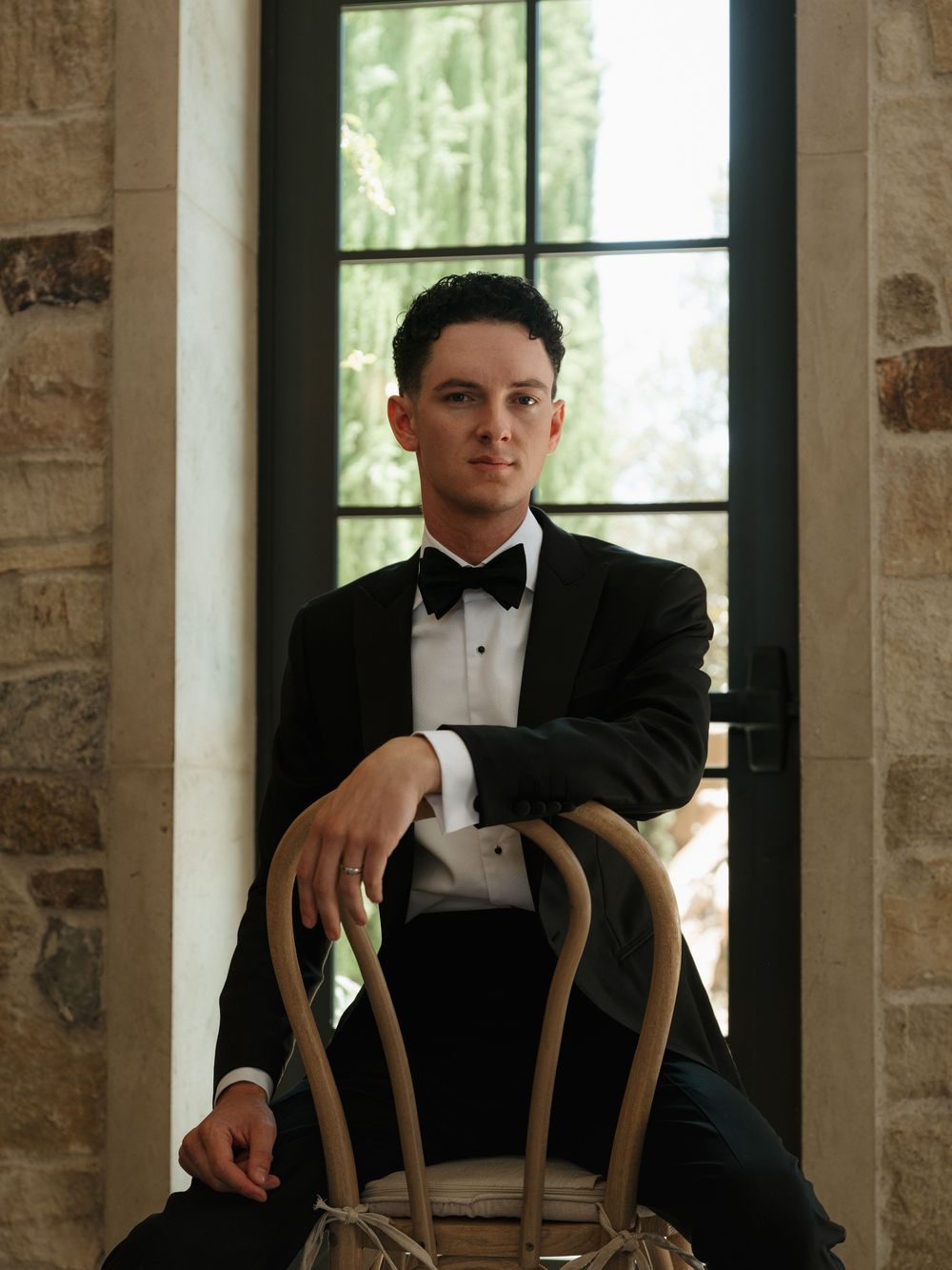 Young man in black tuxedo and bow tie sitting on wooden chair by stone wall window.