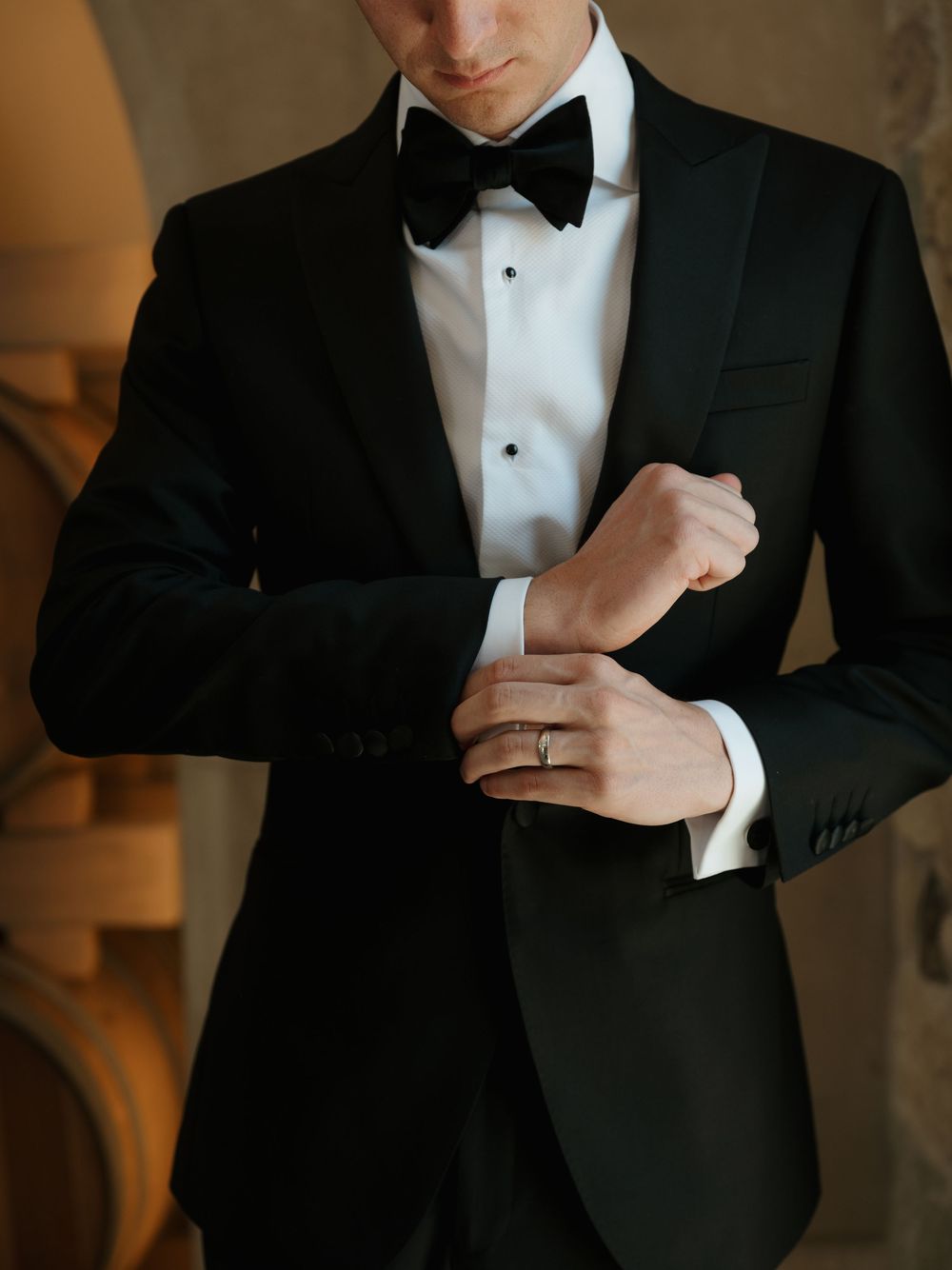 A groom in a black tuxedo with bow tie adjusts his cufflinks, wearing a wedding ring on his finger.