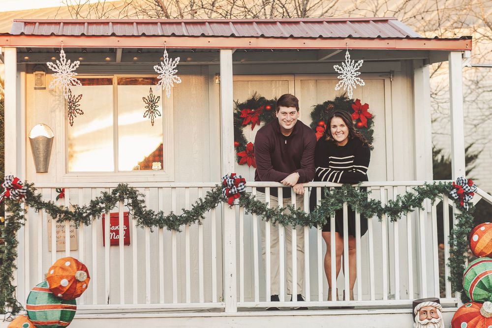 Couple smiling on a festively decorated porch with Christmas garland, wreaths, and colorful ornaments.