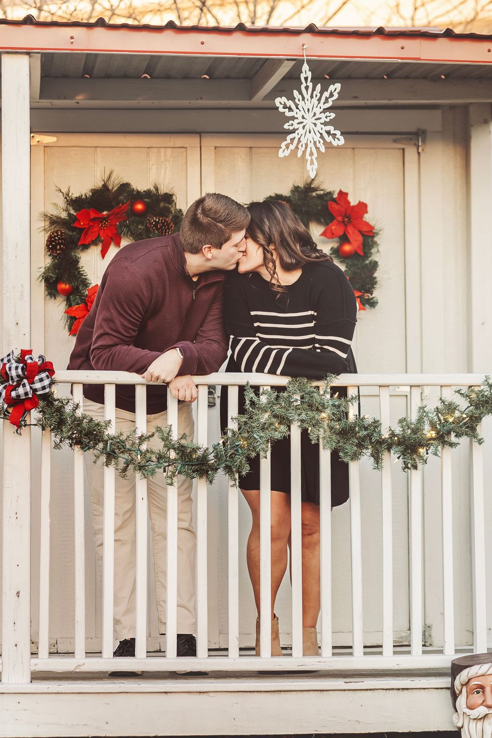 Couple kissing on a Christmas-decorated porch with wreaths, garland, and holiday lights during winter.