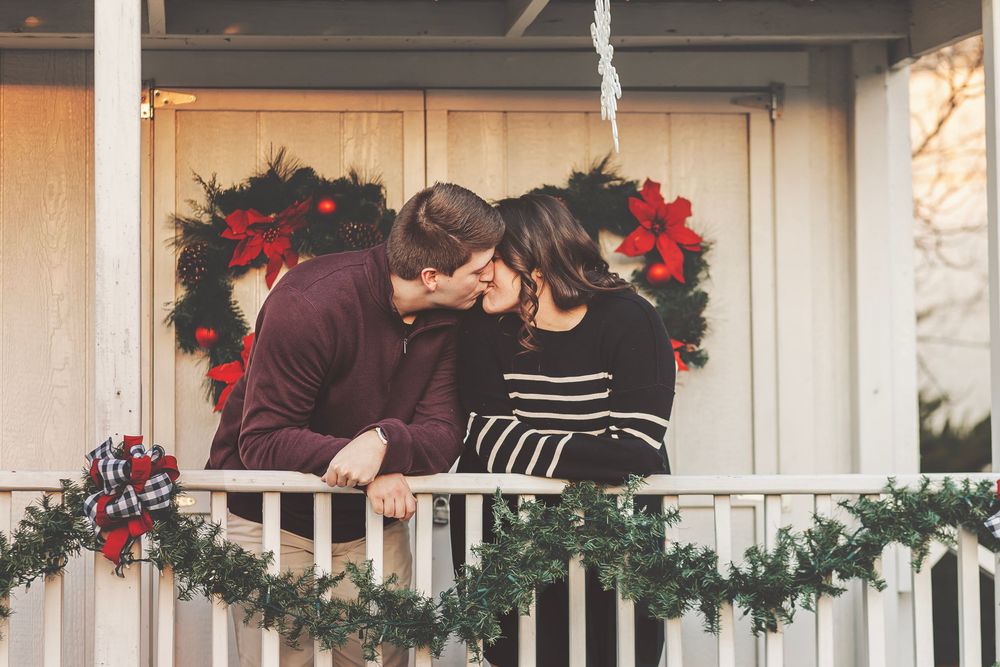 Couple sharing a romantic kiss on a decorated Christmas porch with holiday wreaths and garland.