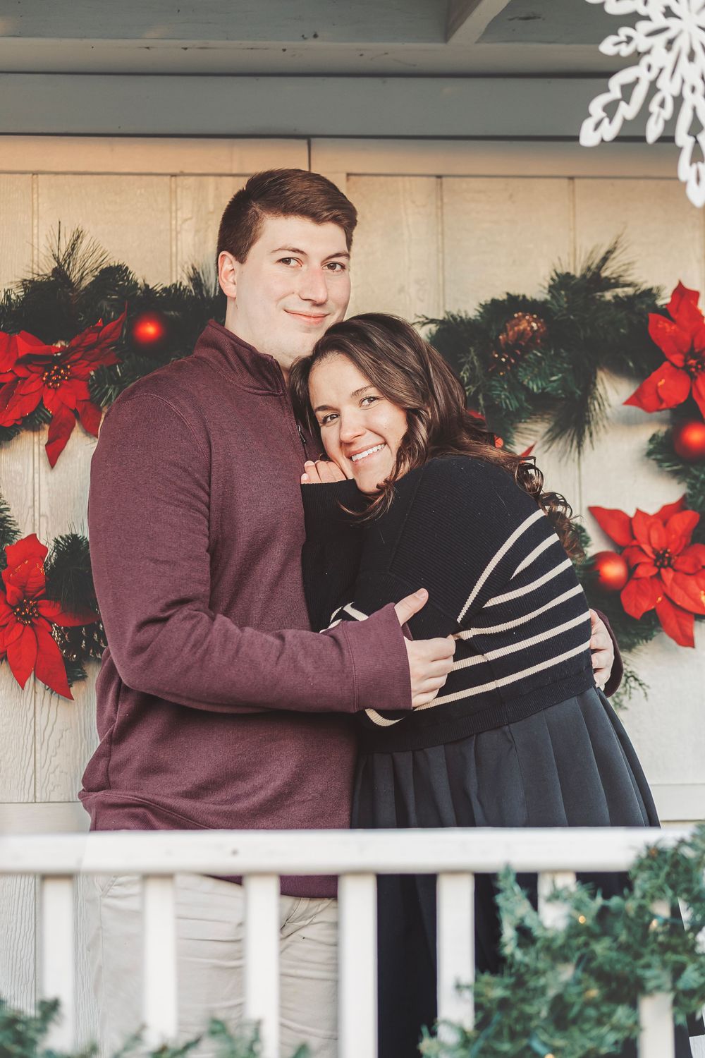 Couple embracing on a decorated Christmas porch with poinsettias and garland, smiling warmly in holiday attire.