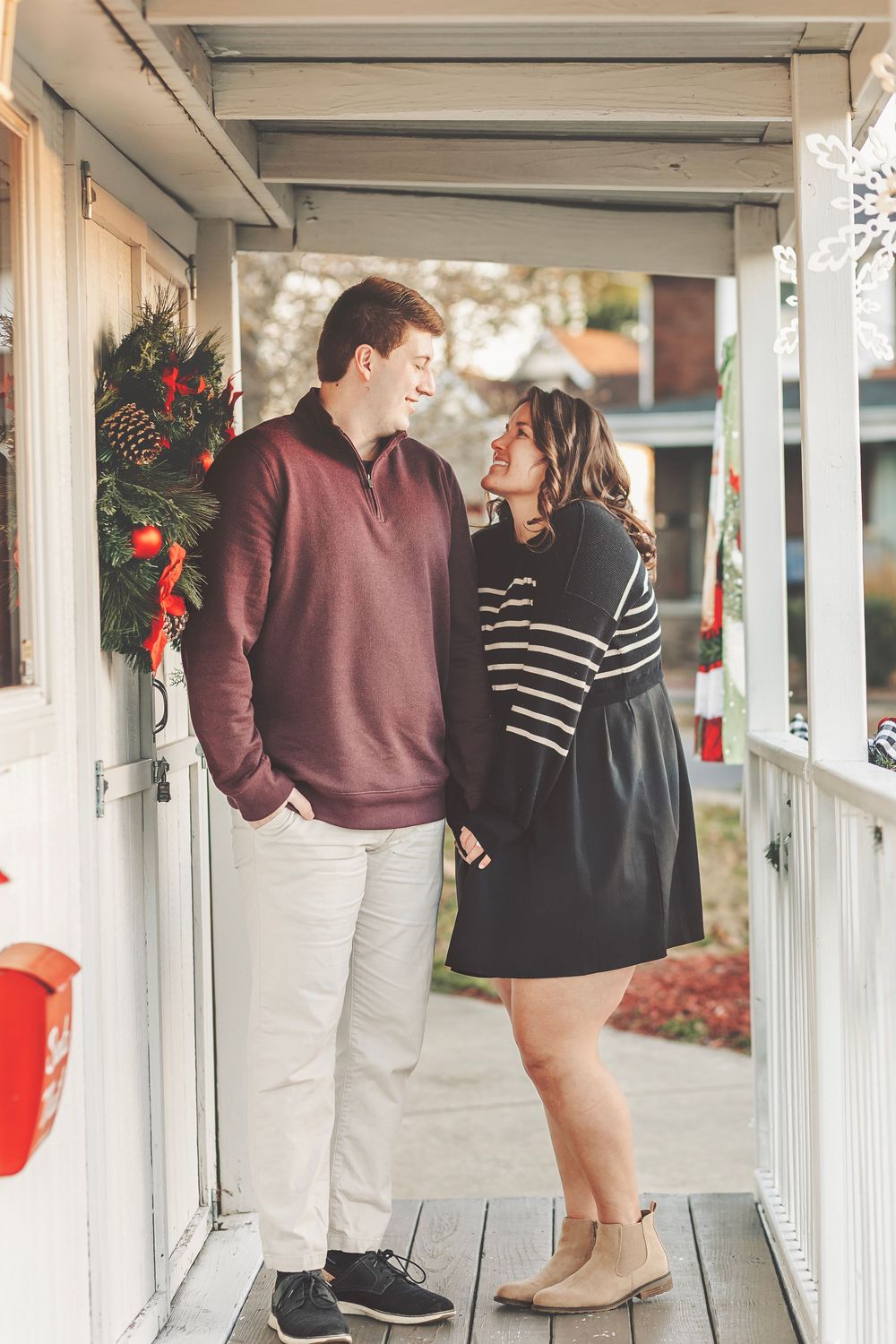Couple sharing a romantic moment on a decorated Christmas porch, woman in striped top and man in burgundy sweater.