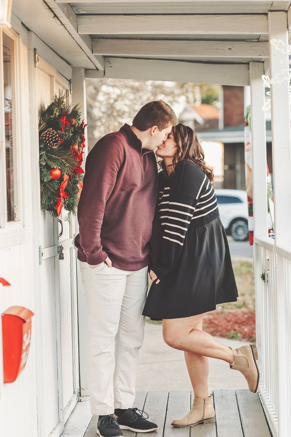 Couple sharing a romantic kiss on a decorated Christmas porch, woman in striped top and black skirt, man in burgundy sweater.